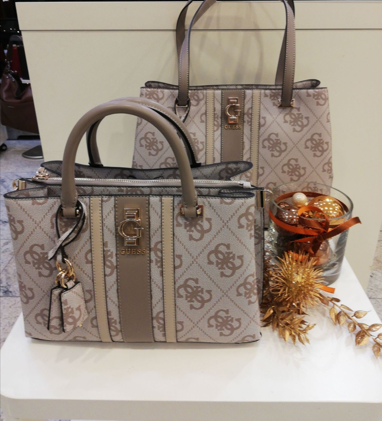 Zwei Guess-Handtaschen mit braunen und weißen Logos auf einem weißen Tisch, eine mit einer Reißverschlusstasche und einem Schlüsselanhänger, die andere mit einem braunen Griff und einem goldenen Logo.