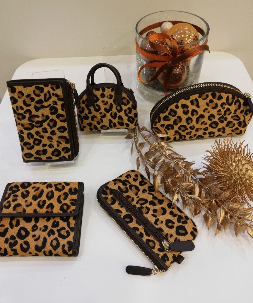 Eine Sammlung von Leopardenprint-Lederaccessoires auf einem weißen Tisch, einschließlich einer Geldbörse, Handtaschen und einer kleinen Handtasche. Eine Glasschale mit goldenen Ornamenten und Bändern verleiht dem Ganzen eine dekorative Note.