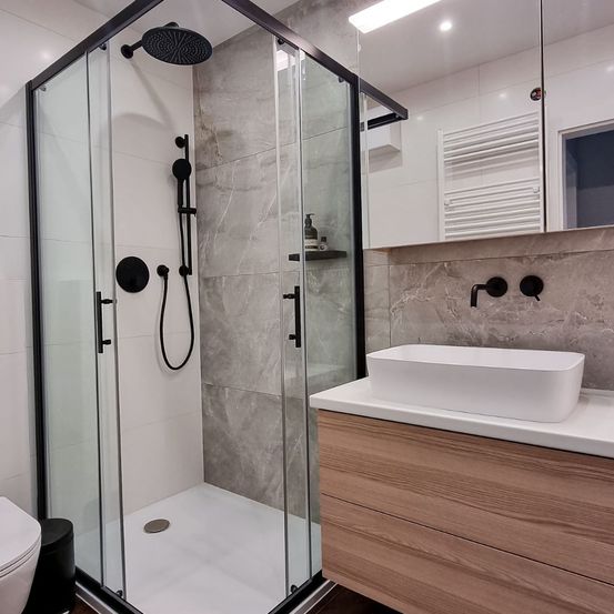 Ein modernes Badezimmer mit einer begehbaren Dusche, einem weißen Waschbecken und einem schwarzen Toilettensitz. Graue Fliesen und Holzschränke verleihen dem Raum Eleganz.