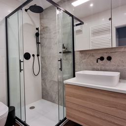 Ein modernes Badezimmer mit einer begehbaren Dusche, einem weißen Waschbecken und einem schwarzen Toilettensitz. Graue Fliesen und Holzschränke verleihen dem Raum Eleganz.