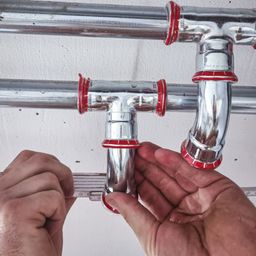 Eine Person misst und justiert eine silberne Rohrleitung mit roten Verschlüssen an einer weißen Wand.