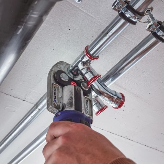 Eine Nahaufnahme einer Person, die ein blaues und silbernes Werkzeug benutzt, um an Metallrohren zu arbeiten, die an einer weißen Decke befestigt sind.