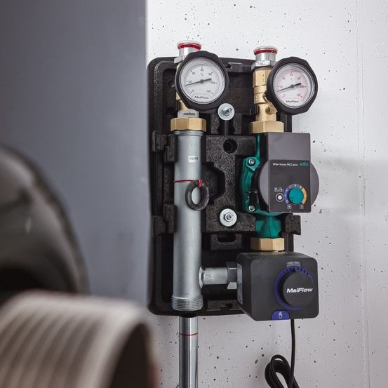 Ein schwarzes Montagegehäuse enthält verschiedene Komponenten wie Manometer, Ventile und Zähler. Die Geräte sind an einem Stahlstab angeschlossen. Es ist an einer weißen Wand montiert.