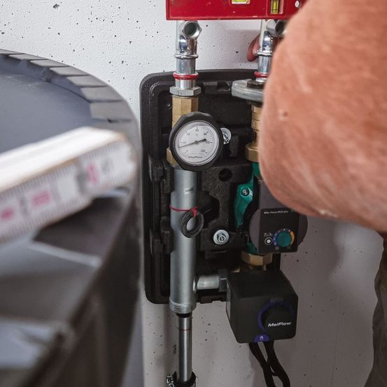 Eine Person arbeitet an einer wandmontierten Wasseranlage, wobei ein Messgerät zur Druckmessung verwendet wird und ein Ventil eingestellt wird. In der Nähe sind ein Messband und ein schwarzer zylindrischer Gegenstand zu sehen.