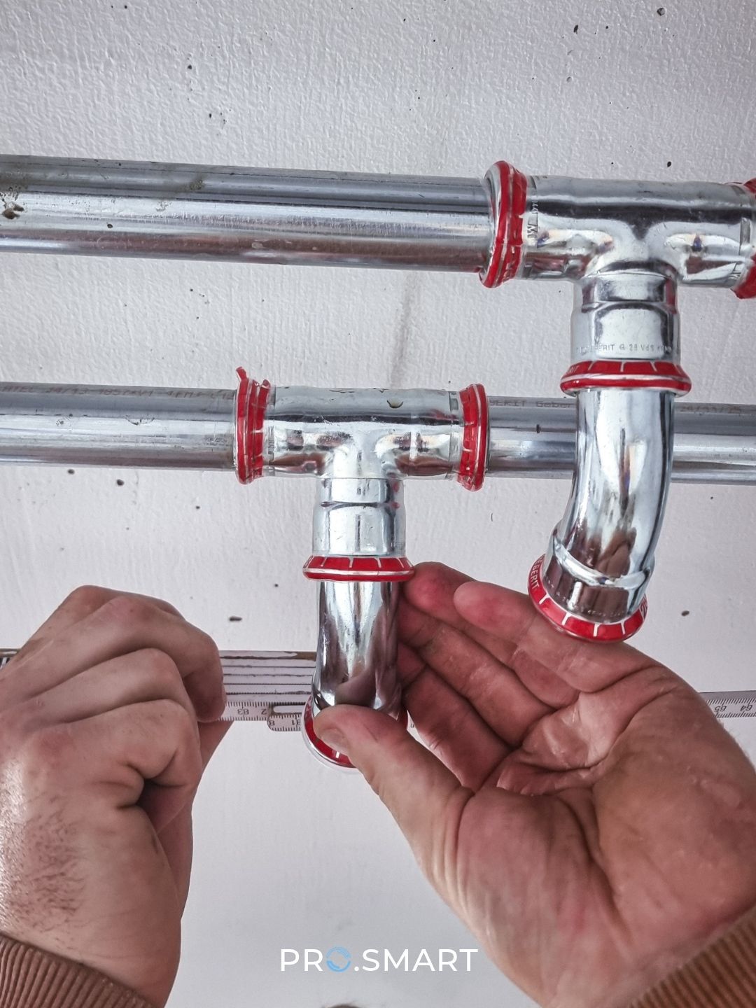 Eine Person misst eine silberne Rohrleitung mit rotem Kappe mit einem Messband. Das Rohr ist an einer weißen Wand montiert.