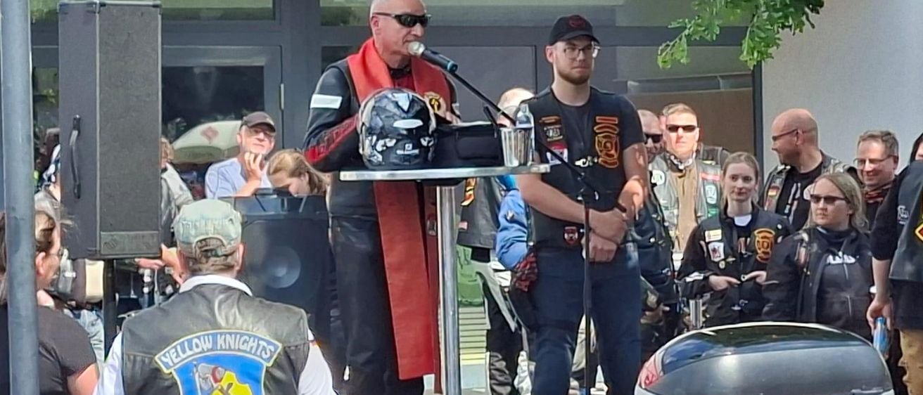 Ein Mann steht an einem Rednerpult mit Mikrofon, trägt einen roten Schal und eine Lederweste mit dem Yellow Knights Germany Logo. Neben ihm steht ein Motorradhelm auf einem Tisch. Im Hintergrund beobachtet eine Gruppe von Menschen, einige von ihnen tragen Hüte und Sonnenbrillen.