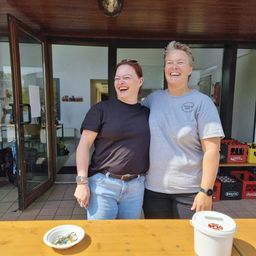 Zwei Frauen stehen lächelnd zusammen vor einem Holztisch mit einem Eimer und einer Schüssel. Sie befinden sich in der Nähe einer offenen Tür, die zu einem Flur führt.