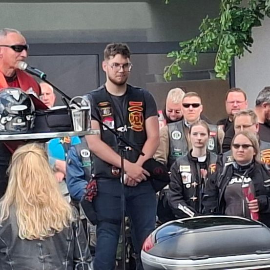 Eine Gruppe von Menschen steht um ein Podium herum, an dem ein Mann in ein Mikrofon spricht. Eine Frau mit langen blonden Haaren ist im Vordergrund zu sehen, und ein Helm ruht auf einem Tisch in der Nähe. Der Mann in der Mitte trägt ein schwarzes T-Shirt und eine Lederweste.