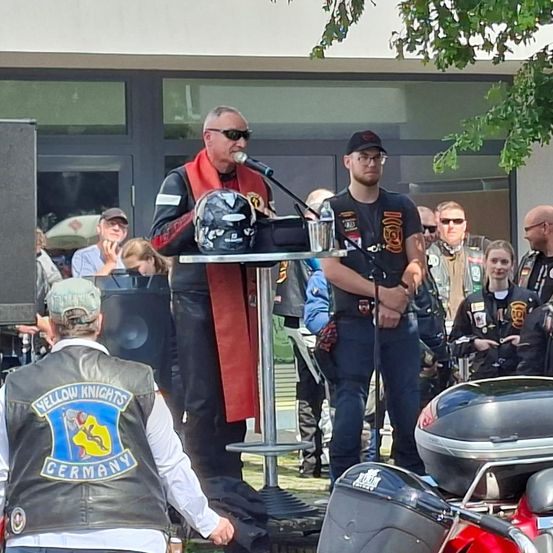 Ein Mann mit rotem Schal spricht in ein Mikrofon. Mehrere Menschen stehen um ihn herum, darunter ein Mann mit Mütze und eine Frau. Ein Motorradhelm liegt auf einem Tisch.