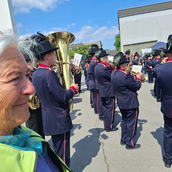 Eine Gruppe von Musikern in dunkelblauen Uniformen mit roten und weißen Streifen spielt auf einer Straße, während eine Frau von links zuschaut.