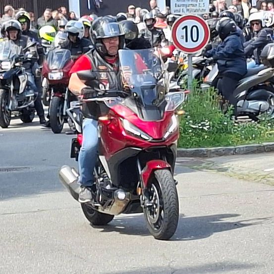 Eine Gruppe von Motorradfahrern mit Helmen fährt auf der Straße. Ein Mann in einem roten T-Shirt und blauen Jeans sitzt vorne. Ein Geschwindigkeitsschild steht am Straßenrand.