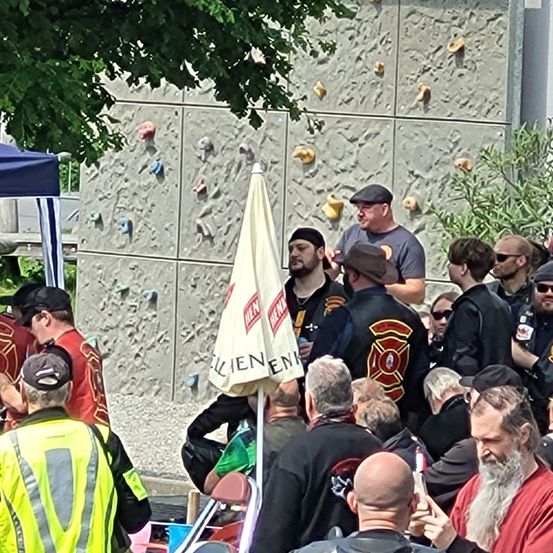 Eine Gruppe von Männern, einige in Motorradjacken, steht um einen geöffneten Regenschirm herum. Dahinter ist eine Kletterwand zu sehen. Ein Mann hält ein Handy.