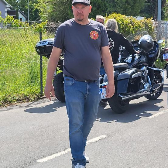 Ein Mann geht eine Straße entlang, hinter ihm steht ein Motorrad. Er trägt ein graues Shirt mit einem Feuerwehrlogo. Im Hintergrund steht eine Frau neben dem Motorrad.