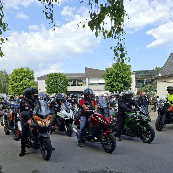 Viele Motorradfahrer versammeln sich auf einem Parkplatz. Einige tragen Westen und Helme. Bäume und Gebäude sind im Hintergrund. Der Himmel ist teilweise bewölkt.