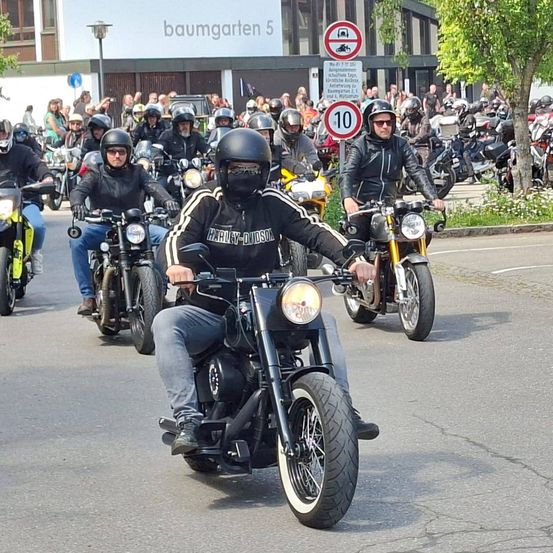 Motorradfahrer fahren in einer Parade; einige tragen Helme, Jacken und Handschuhe. Ein Gebäude mit 'baumgarten 5' ist im Hintergrund.