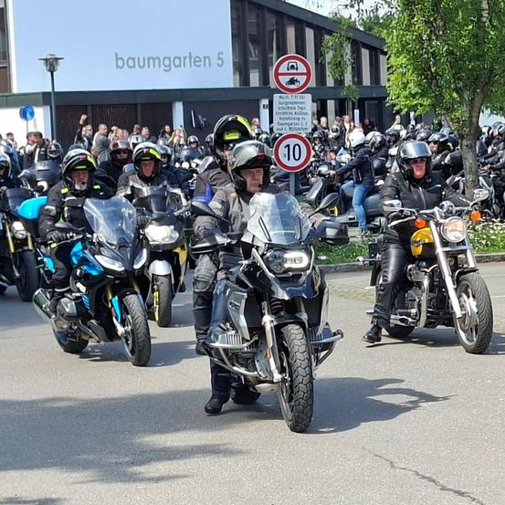 Eine Gruppe von Motorradfahrern fährt auf einer Straße vor einem Gebäude mit dem Schild 'baumgarten 5'. Sie tragen Helme und Lederjacken. Dahinter stehen Menschen und beobachten. Im Hintergrund befinden sich zwei Verkehrsschilder.