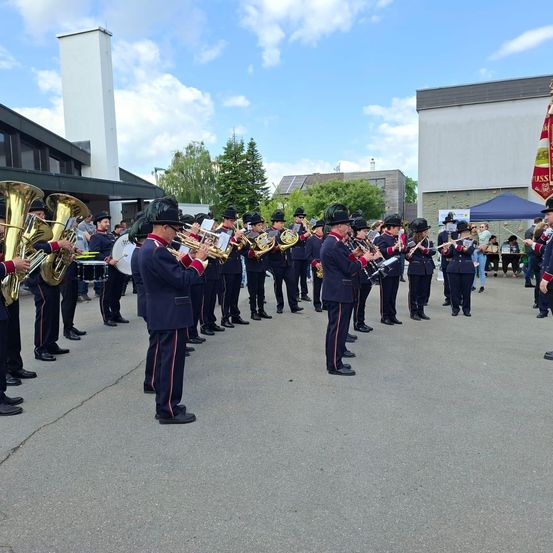 Eine Blaskapelle, gekleidet in marineblaue Uniformen, spielt im Freien. Sie spielen eine Vielzahl von Instrumenten, darunter Trompeten, Posaunen und Trommeln. Der Ort ist ein gepflasterter Bereich mit Gebäuden und Bäumen im Hintergrund.