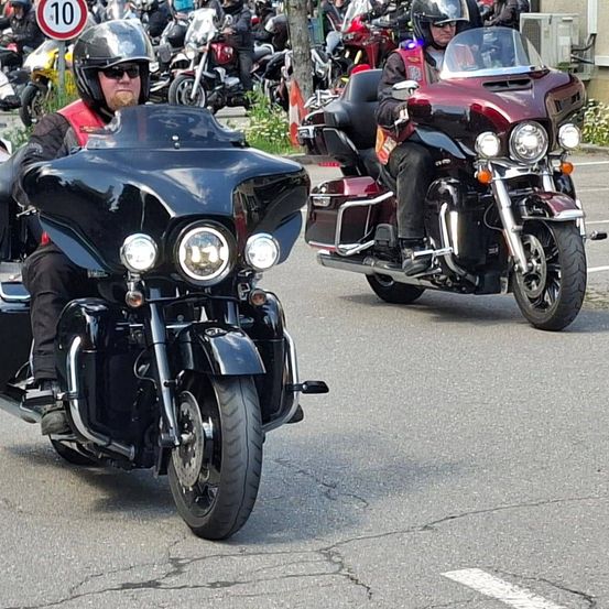 Eine Gruppe von Motorradfahrern fährt auf der Straße. Ein Mann auf einem schwarzen Motorrad führt den Weg, gefolgt von einem weiteren Fahrer auf einem roten Motorrad.
