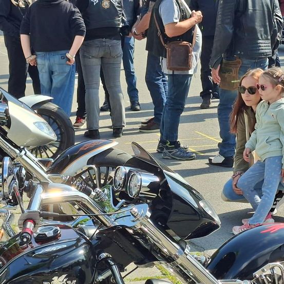 Mehrere Menschen stehen neben Motorrädern, darunter ein Mann mit einer braunen Tasche, eine Frau mit Sonnenbrille und ein Kind mit rosa Schuhen. Sie scheinen auf einem Motorradtreffen zu sein.