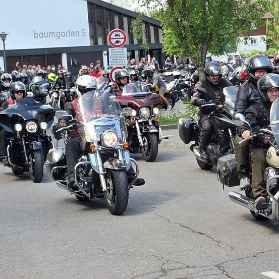 Eine Gruppe von Motorradfahrern fährt auf der Straße, vorbei an einem Gebäude mit dem Schild 'baumgarten 5'. Viele Menschen stehen hinter einem Zaun und beobachten die Veranstaltung. Die Motorräder sind entlang der Straße geparkt.