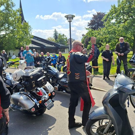 Eine Gruppe von Menschen versammelt sich um geparkte Motorräder, ein Mann winkt, während eine Frau einen Becher hält. Bäume und ein Gebäude sind im Hintergrund.