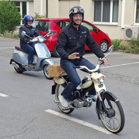 Zwei Motorradfahrer fahren auf einer Straße, einer mit einem Aufbewahrungsbox. Ein rotes Auto ist dahinter geparkt. Der Fahrer vorne trägt Helm und Schutzbrille.