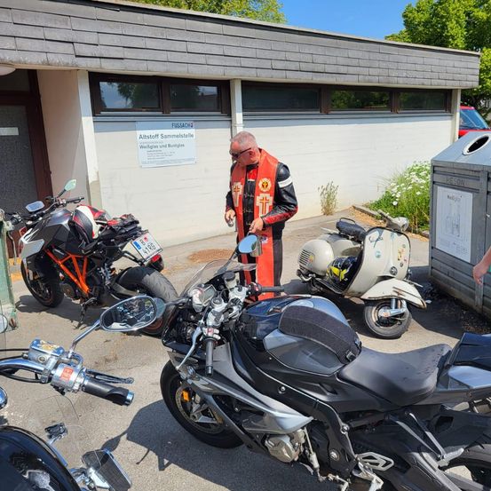 Ein Mann in einem roten Weste steht neben einem schwarzen Motorrad, mit anderen Motorrädern in der Nähe. Ein Gebäude mit einem Schild, das 'Altstoff Sammelstelle' liest, ist hinter ihm.