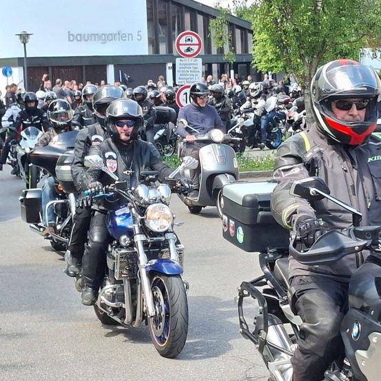 Eine große Gruppe von Motorradfahrern in Helmen und Motorradkleidung fährt mit ihren Motorrädern in einer Parade. Im Hintergrund ist ein Schild mit der Nummer 1 und einem Parkverbotsschild zu sehen.