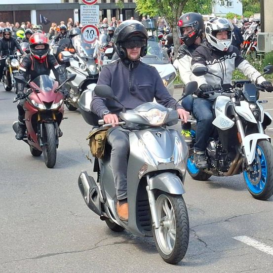 Eine Gruppe von Motorradfahrern fährt auf einer Straße, an der sich eine Menge Schaulustiger versammelt hat. Die Fahrer tragen Helme und Schutzkleidung. Geschwindigkeitsbegrenzungsschilder sind sichtbar.