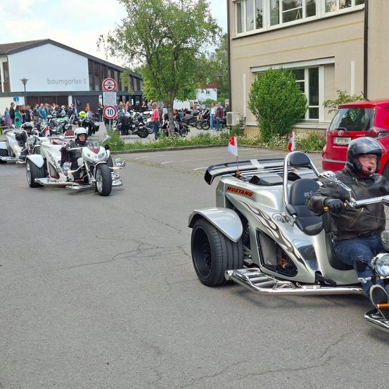 Motorräder stehen auf einer Straße in einer Reihe. Ein Mann mit Helm fährt vor einer Menge und Gebäuden. Ein rotes Auto ist in der Nähe geparkt.