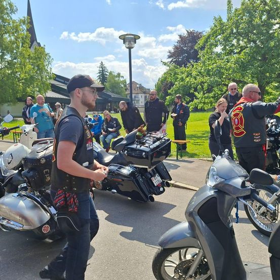 Eine Gruppe von Menschen versammelt sich in der Nähe von Motorrädern, einige sitzen auf dem Boden, andere stehen, unter einem klaren Himmel mit Bäumen im Hintergrund.