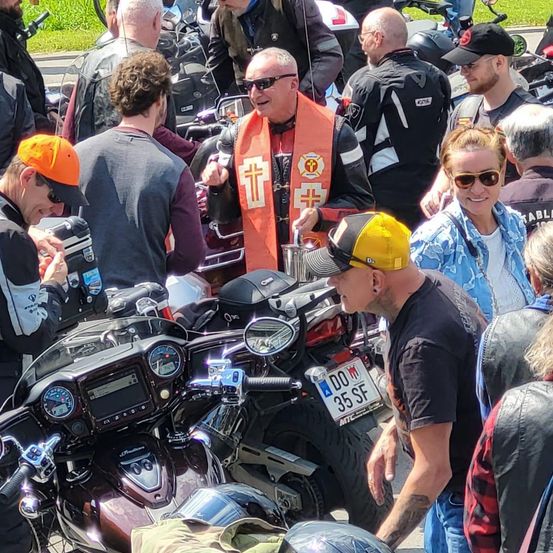 Eine Gruppe von Motorradfahrern versammelt sich an einem sonnigen Tag. Ein Mann in einer orangefarbenen Weste mit einem Kreuz spricht mit einem anderen Mann in einer gelben Mütze. Andere Fahrer in Lederjacken und Sonnenbrillen umgeben sie.