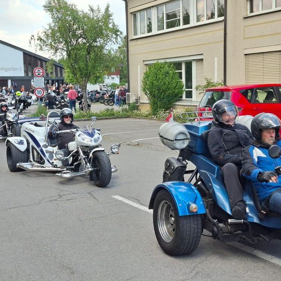 Eine Gruppe von Motorradfahrern fährt in einer Parade, alle tragen Helme. Einige sind auf blauen Dreirädern, andere auf Motorrädern. Dahinter ist ein rotes Auto geparkt und im Hintergrund steht ein Gebäude.