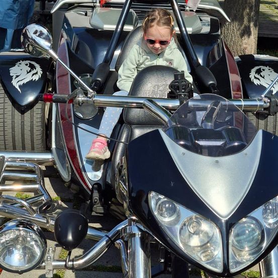 Ein kleines Mädchen mit Sonnenbrille sitzt in einem schwarzen und roten Dreiradmotorrad mit Löwendesign an den Seiten.