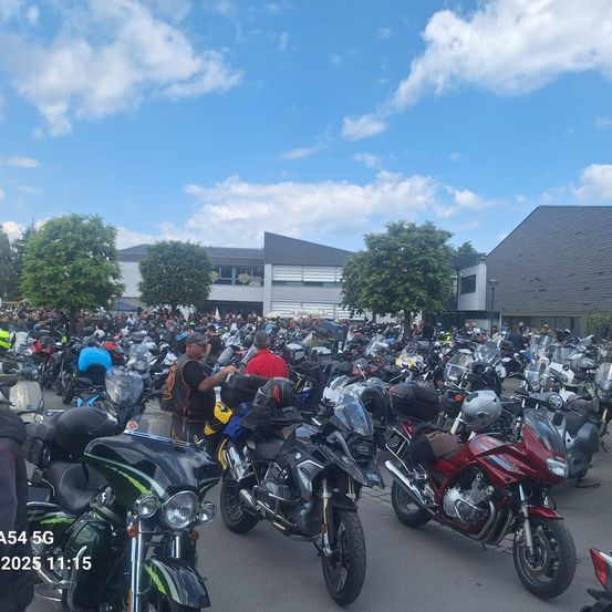 Eine große Gruppe von Motorrädern ist auf einem Parkplatz vor einem Gebäude unter einem blauen Himmel mit Wolken geparkt.