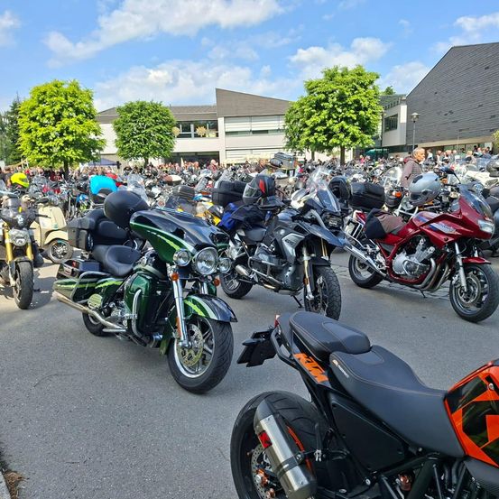 Eine große Ansammlung von Motorrädern im Freien vor einem Gebäude mit Bäumen und Menschen in der Umgebung.