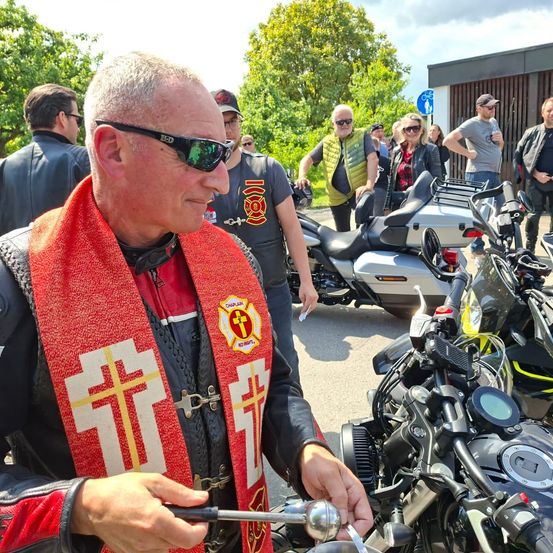 Ein Mann mit einem roten Schal mit Kreuzen steht vor einem Motorrad, möglicherweise bereit, einen Segen zu sprechen. Hinter ihm versammeln sich mehrere Menschen, und Motorräder sind geparkt.
