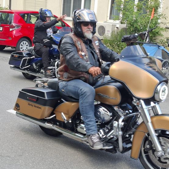 Zwei Motorradfahrer fahren die Straße entlang, einer auf einem blauen Motorrad und der andere auf einem goldenen Harley-Davidson. Ein rotes Auto und ein Gebäude sind im Hintergrund zu sehen.