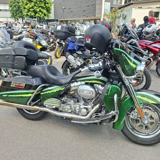 Ein grünes Harley Davidson Motorrad steht in einem überfüllten Parkplatz, umgeben von anderen Motorrädern. Die Gegend ist voller Menschen, und ein Gebäude ist im Hintergrund zu sehen.