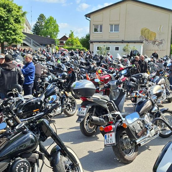 Eine große Menschenmenge und Motorräder versammeln sich vor einem Gebäude mit einem Wandbild. Einige Leute stehen, andere sitzen auf ihren Motorrädern. Das Gebäude hat mehrere Fenster und eine Treppe.