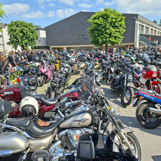 Eine große Ansammlung von Motorrädern ist vor einem Gebäude geparkt, vor dem sich viele Menschen aufhalten. Im Hintergrund sind Bäume und ein bewölkter Himmel zu sehen.