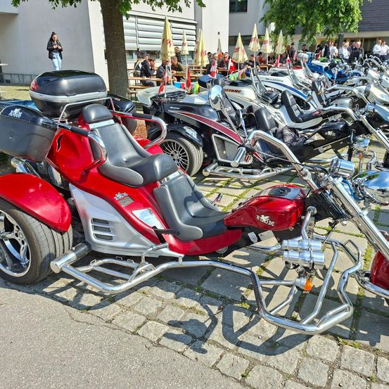 Ein roter Dreirad-Motorroller mit schwarzem Sitz steht auf einer Kopfsteinpflasterstraße, mit mehreren anderen Motorrädern dahinter. Ein Gebäude mit vielen Menschen ist im Hintergrund.