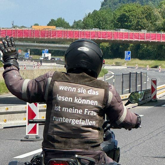 Ein Motorradfahrer mit schwarzem Helm winkt auf der Straße, mit einer Brücke im Hintergrund. Sein Vest sagt: 'Wenn Sie das lesen können, ist meine Frau runtergefallen.'