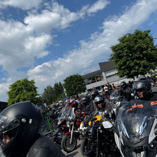 Eine Gruppe von Motorradfahrern in voller Ausrüstung wartet auf einem Parkplatz unter einem teilweise bewölkten Himmel. Bäume und ein Gebäude sind im Hintergrund zu sehen.