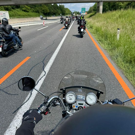 Eine Gruppe von Motorradfahrern fährt auf einer Autobahn mit orangefarbenen Markierungen und einer Überführung darüber. Der Fahrer im Vordergrund trägt Helm und Handschuhe.