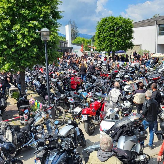 Eine große Menschenmenge von Motorradfahrern hat sich auf einem Parkplatz versammelt, wo zahlreiche Motorräder geparkt sind. Leute gehen herum, einige tragen Helme. Im Hintergrund sind Bäume und Gebäude zu sehen.