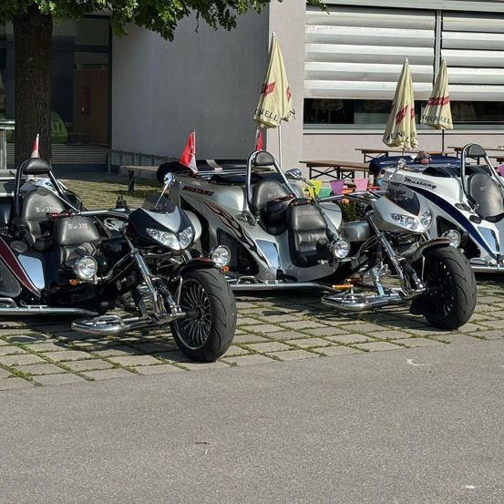 Drei Motorräder sind vor einem Gebäude geparkt. Zwei davon sind Dreiräder und eines ist ein normales Motorrad. Jedes hat Flaggen. Dahinter sind Bänke und geschlossene Fenster.
