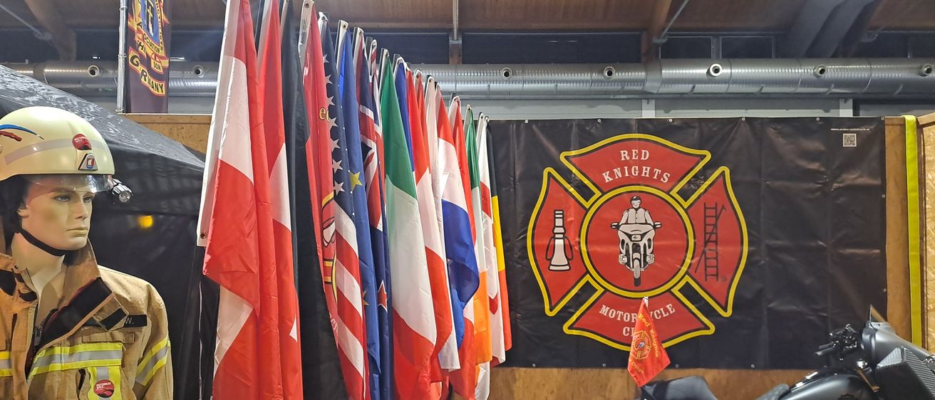 Ein Raum mit Fahnen aus verschiedenen Ländern aufgereiht und einem Motorradclub-Banner an der Wand. Ein Feuerwehrmann-Jacke ist links, und Motorräder sind rechts geparkt.