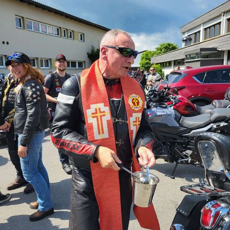 Ein Mann in einer Lederjacke und einem Schal mit Kreuzen hält einen Becher und steht neben einem Motorrad. Dahinter parkt ein rotes Auto. Hinter ihm steht eine Gruppe von Menschen auf einem gepflasterten Bereich.
