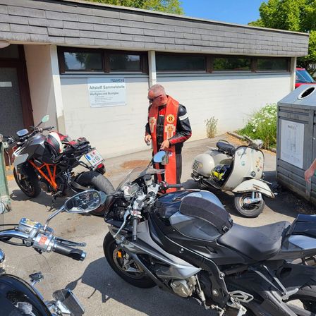 Ein Mann in einem roten Weste steht neben einem schwarzen Motorrad, mit anderen Motorrädern in der Nähe geparkt. Das Gebäude dahinter hat ein Schild, das 'Altstoff Sammelstelle' steht.